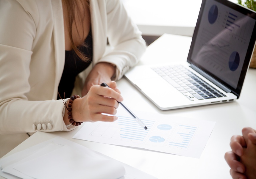 A woman can be seen sitting at a table in front of a laptop with a pen in her hand